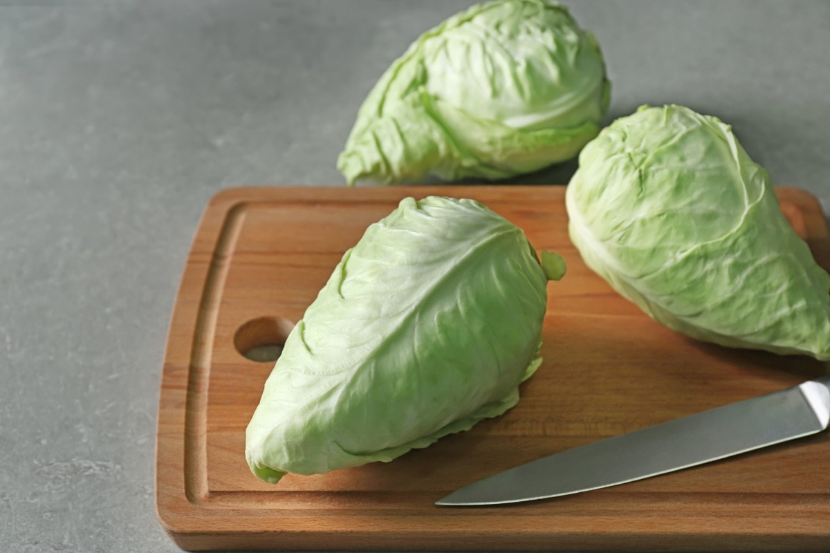 Three pointed cabbages on a wood cutting board next to a kitchen knife.