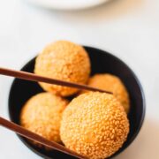 Wooden chopsticks picking up sesame ball from a black bowl on a white table.
