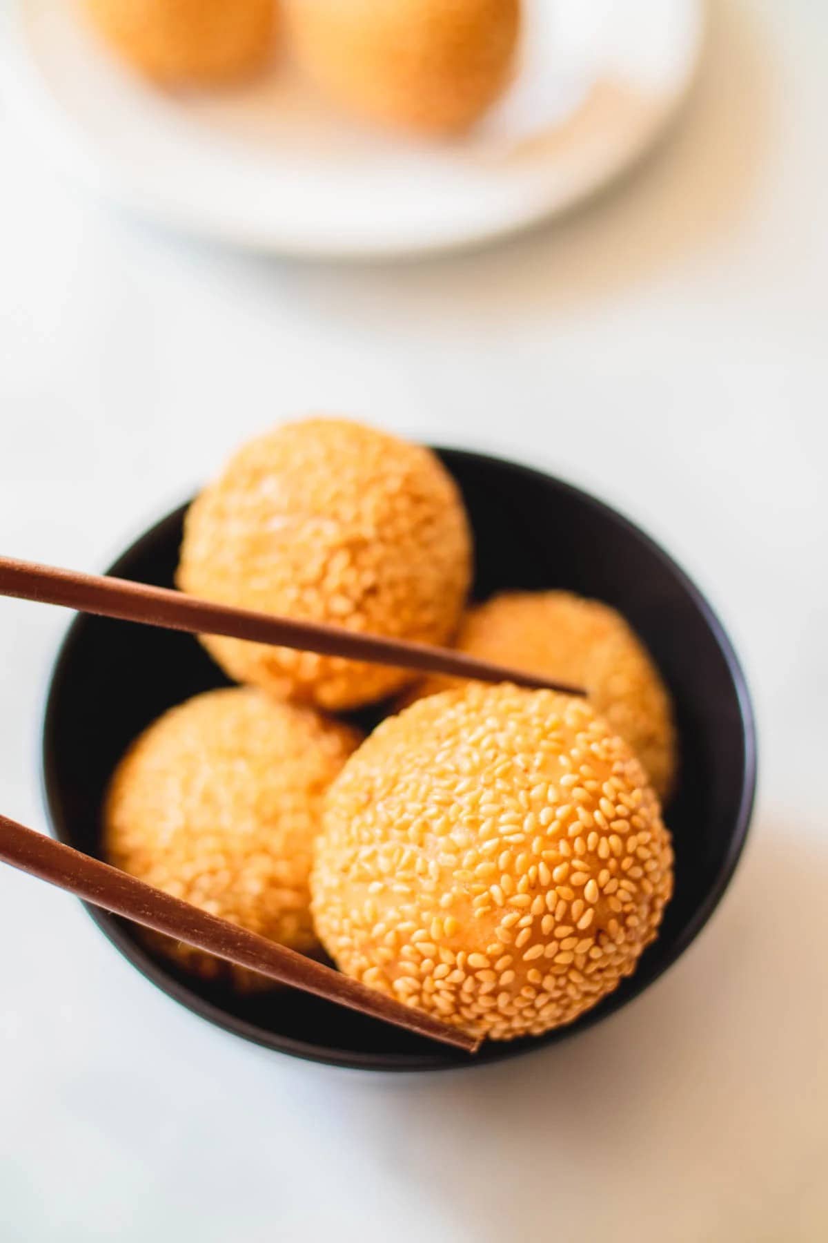 Wooden chopsticks picking up sesame ball from a black bowl on a white table.