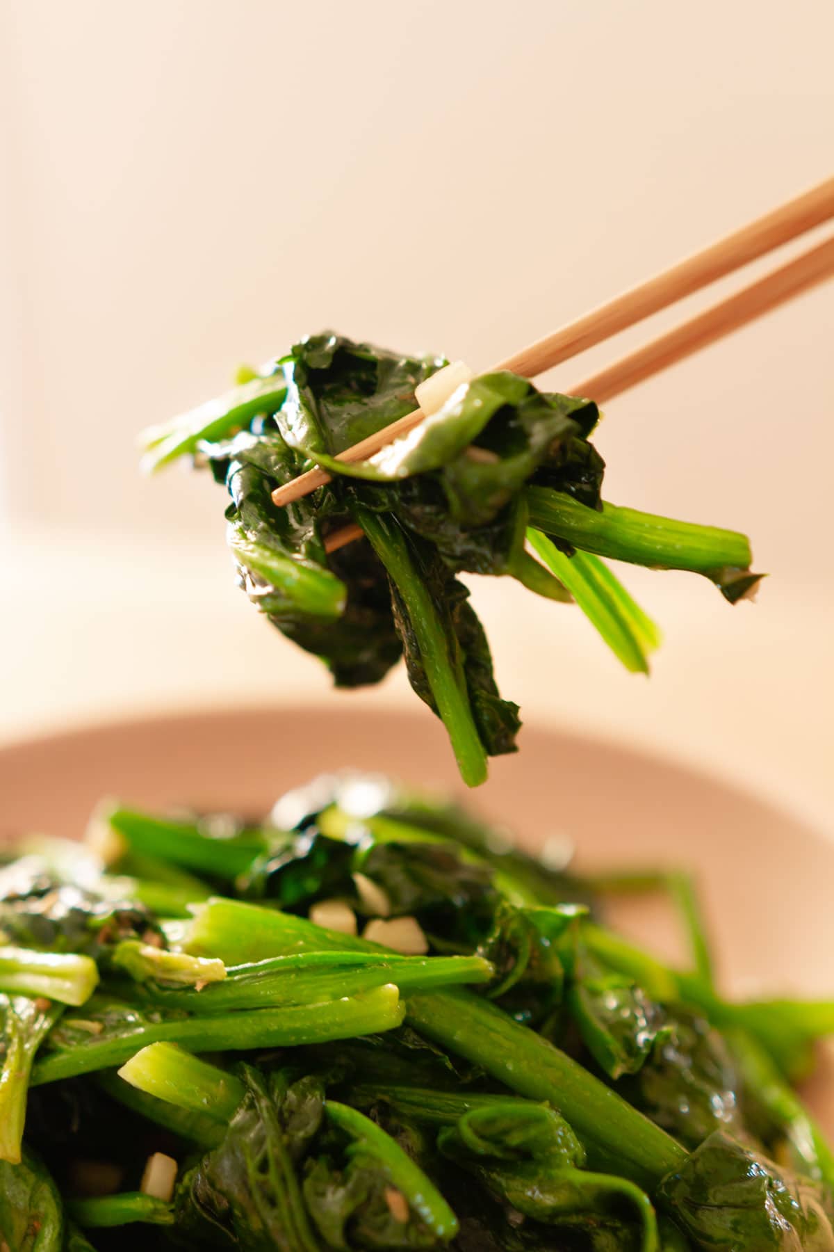 A close up view of chopsticks picking up a bite of stir-fried spinach from a serving plate.