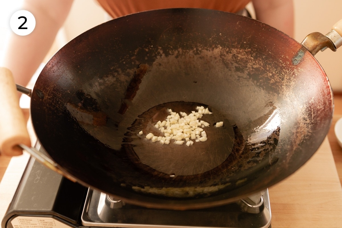 Top down view of a heated wok with canola oil and roughly minced garlic in it, labeled with a circled number "2."