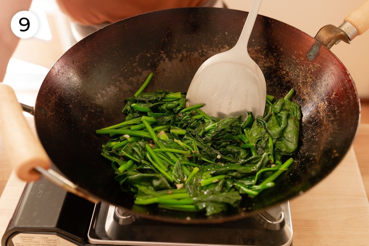 Cindy stirring slightly wilted and bright green spinach in a wok with a metal spatula, labeled with a circled number "9."
