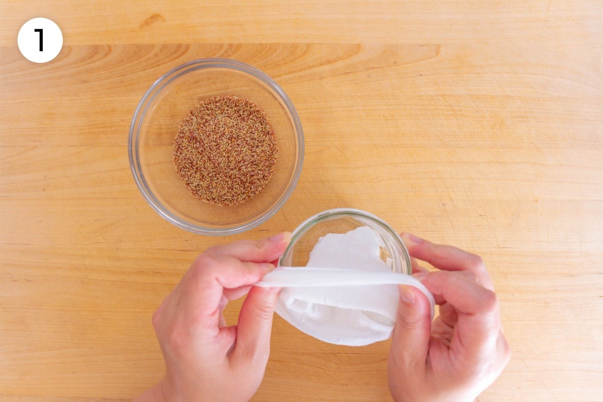 Recipe step 1: Cindy placing the mesh spice bag into a small cup (or mason jar) and wrapping the edges of the bag over the rim of the cup, with a small bowl of aiyu seeds on the side.