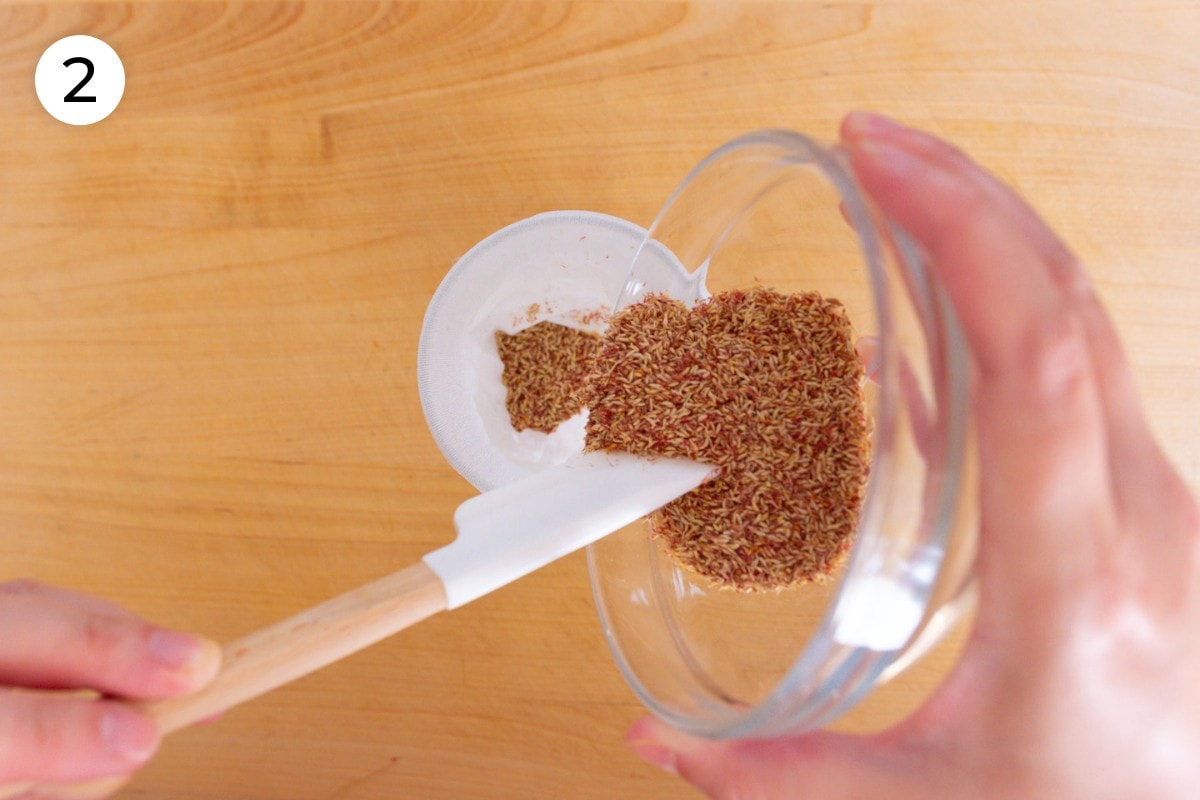 Recipe step 2: Cindy using a small spatula to scoop aiyu seeds into the mesh spice bag that's held open with a small mason jar.