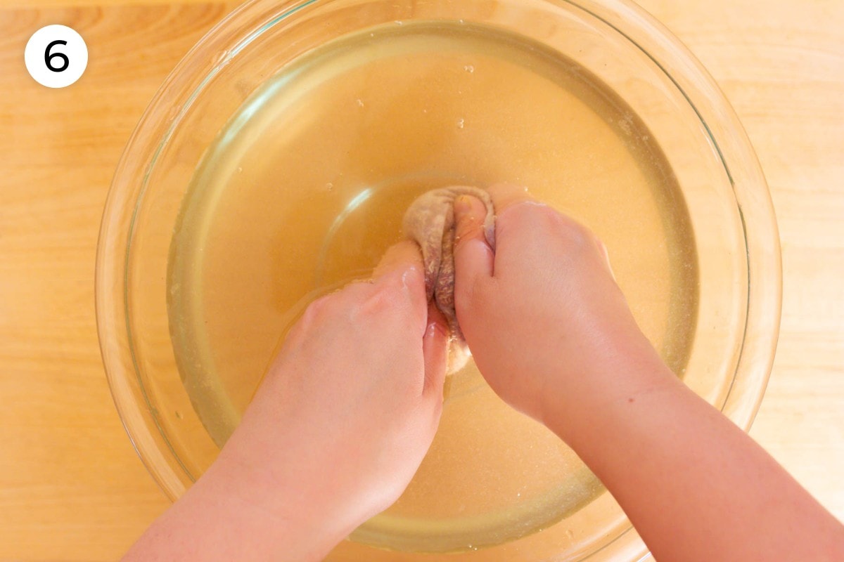 Recipe step 6: Cindy using both hands to rub the aiyu seed filled mesh bag in a large bowl of now translucent pale yellow hard (mineral) water.