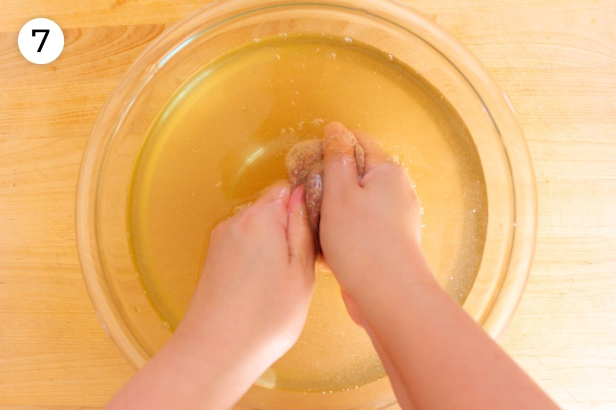 Recipe step 7: Cindy using both hands to rub the aiyu seed filled mesh bag in a large bowl of translucent yellow hard (mineral) water.
