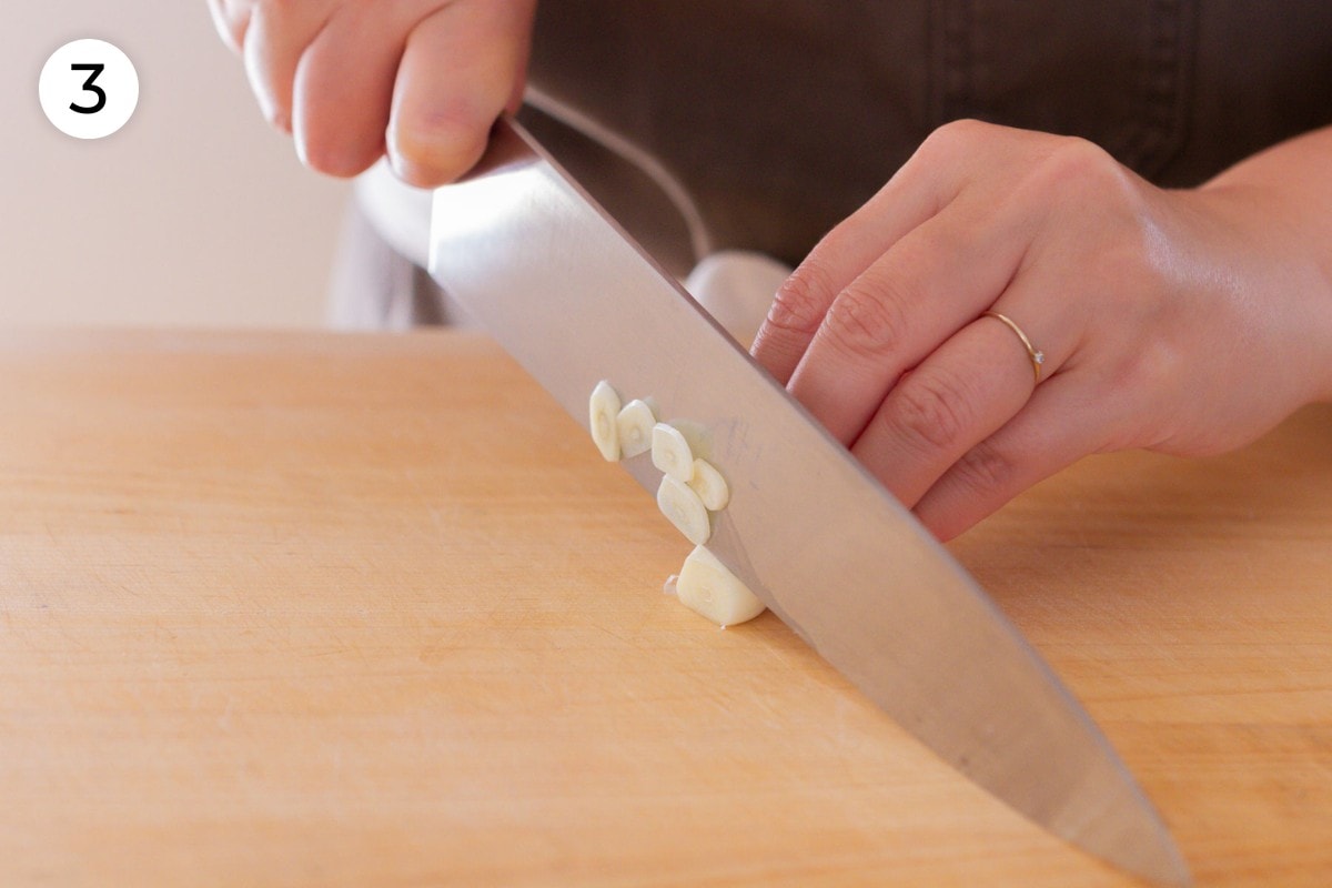 Recipe step 3: Cindy cutting a garlic clove into thin slices with a chefs knife.