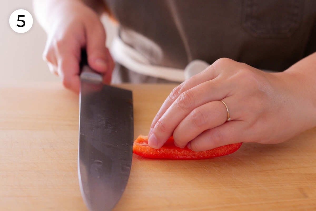 Recipe step 5 (optional): Cindy holding a slice of red bell pepper flat on a cutting board and slicing it with a chefs knife held parallel to the cutting board.