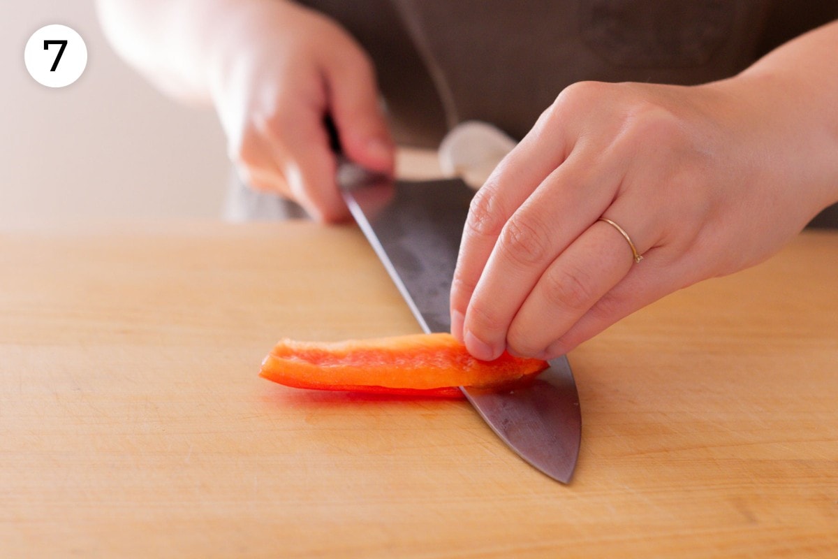 Recipe step 7 (optional): Cindy holding the top of a slice of red bell pepper flat on a cutting board and slicing it close to the skin from left to right with a chefs knife held parallel to the cutting board.