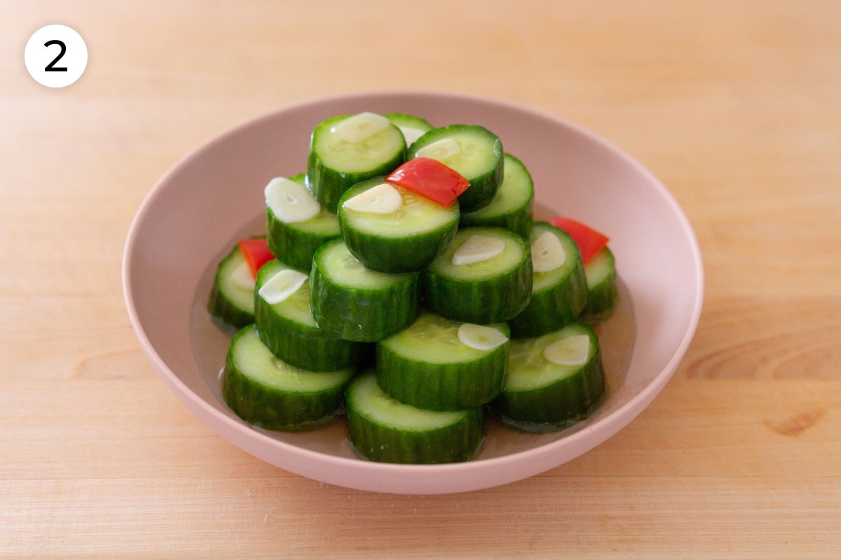 Din Tai Fung cucumber salad recipe plating step 2: Thinly sliced garlic and red chili pepper on top of stacked cucumber slices in a light pink serving plate.