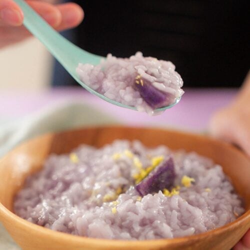 Cindy holding up a spoon full of purple sweet potato congee over a wood bowl filled with congee topped with grated ginger.