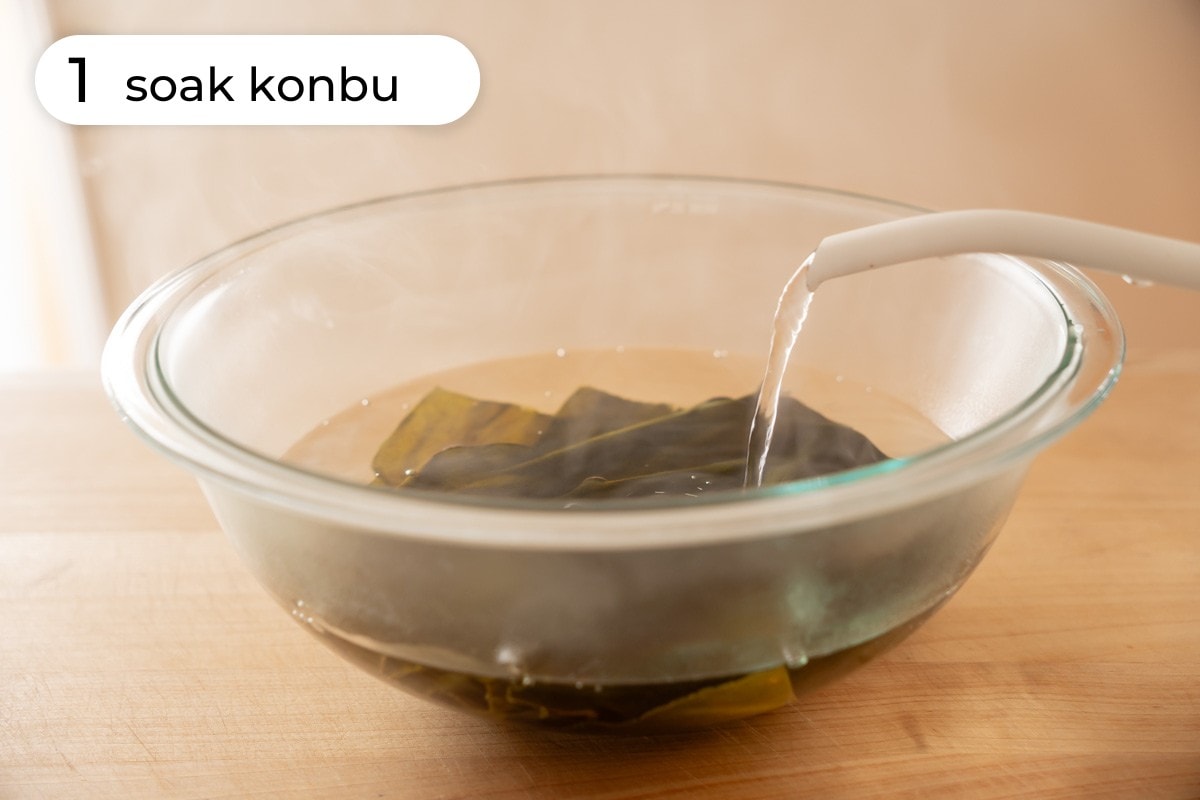 Recipe step 1: Sheets of dry konbu in a large glass mixing bowl being soaked in hot water from a kettle.