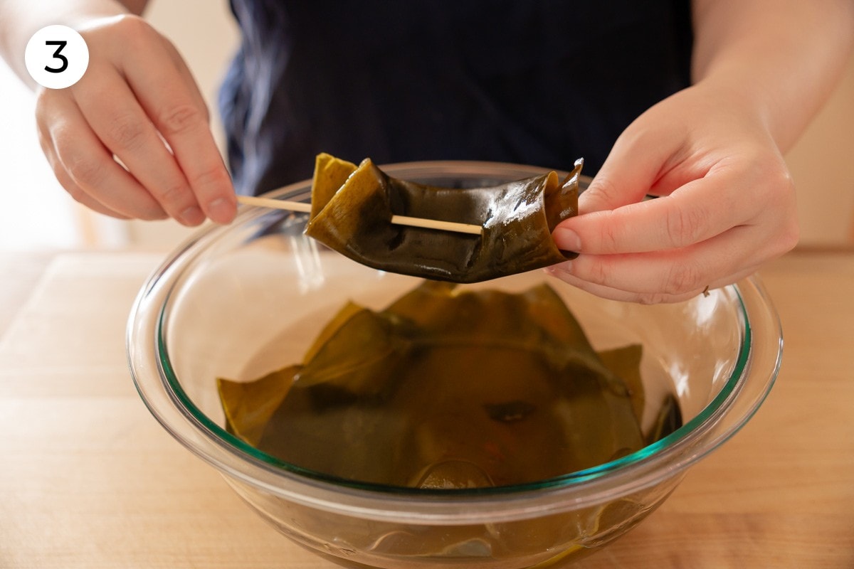 Recipe step 3: Cindy holding up a pre-soaked konbu sheet that has been folded into a 1-inch wide strip and skewered with a toothpick through both ends.
