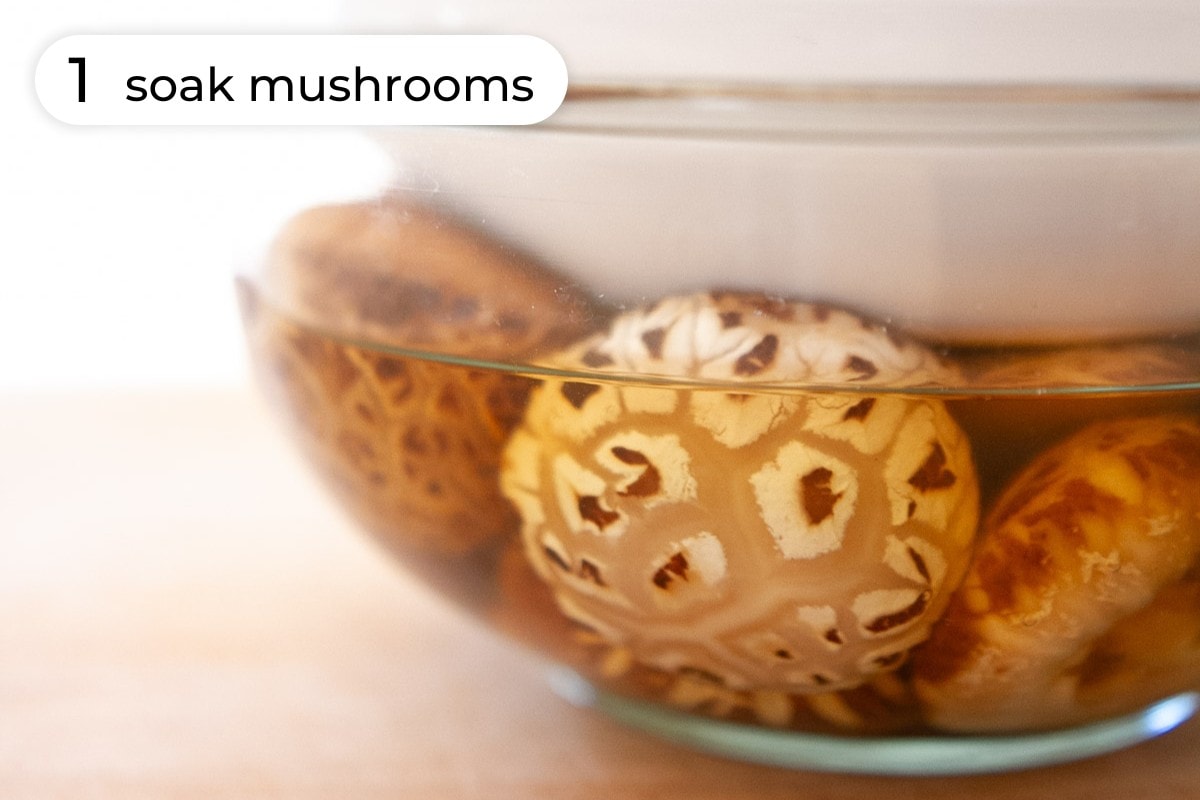 Recipe step 1: Whole dried shiitake mushrooms soaking in water in a large glass bowl with another bowl on top.