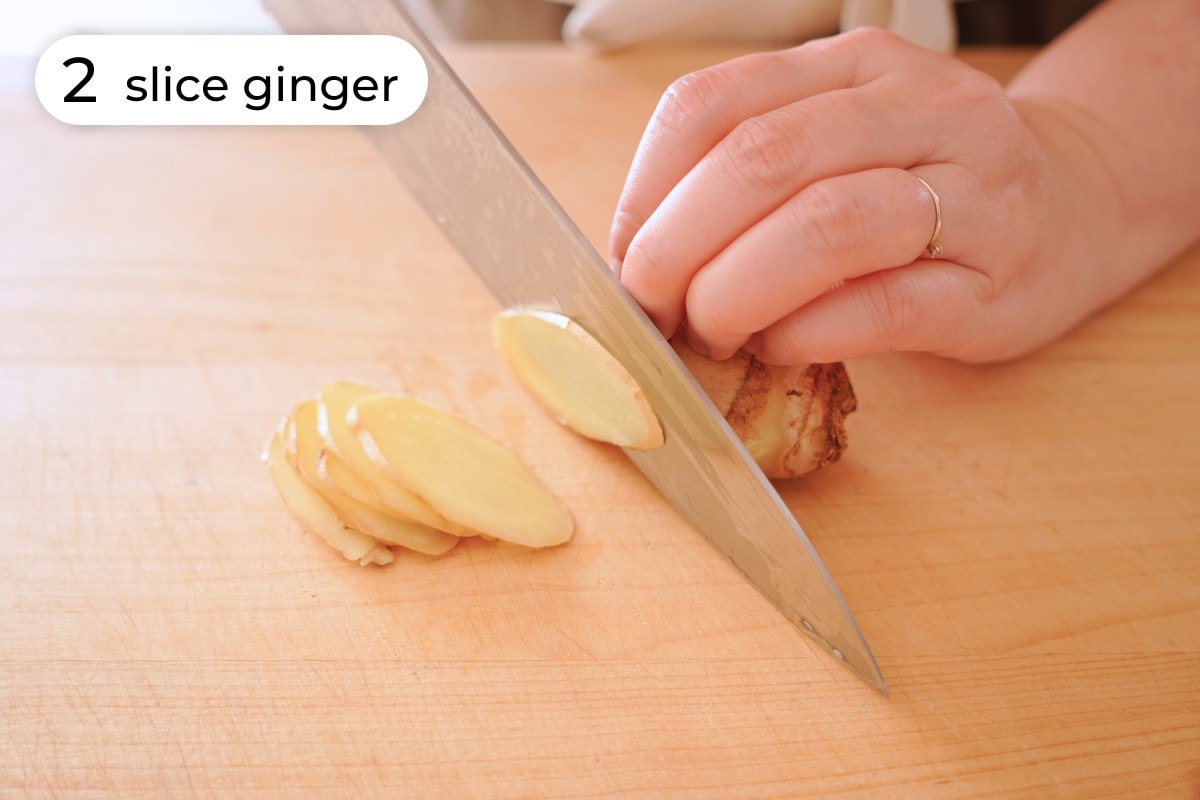 Recipe step 2: Cindy thinly slicing a piece of fresh ginger at an angle with a chefs knife.