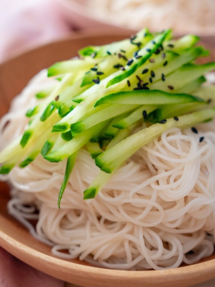 Longevity noodles in a shallow wood bowl, topped with thinly sliced cucumber and toasted black sesame seeds.