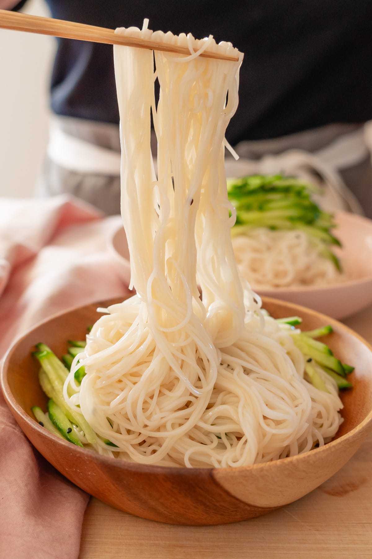 Long noodles being pulled upward from a wood serving bowl with chopsticks.