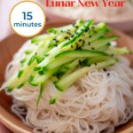 Taiwanese longevity noodles in a shallow wood bowl, topped with thinly sliced cucumber and toasted black sesame seeds and text, "longevity noodles – Lunar New Year," "15 minutes," and "the sound of cooking dot com."