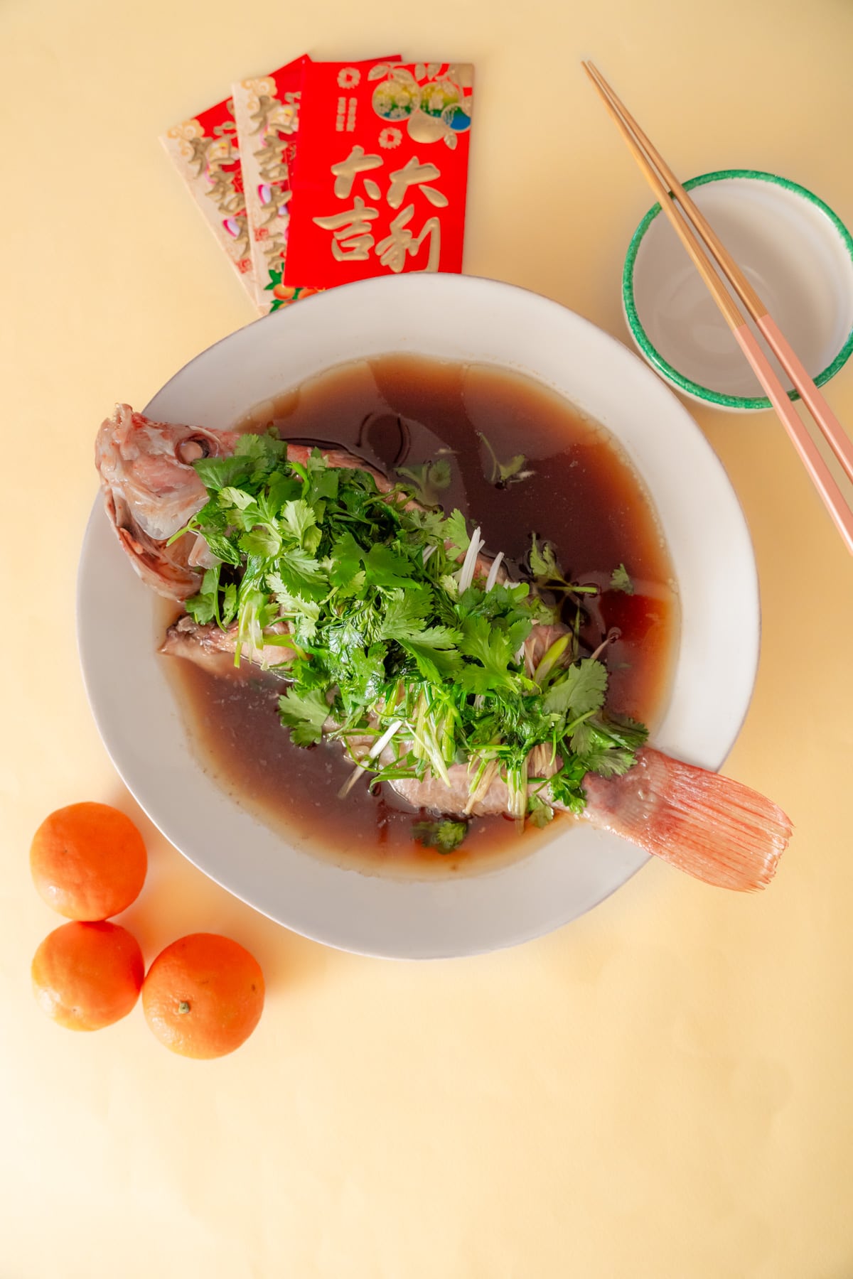 Top-down view of a steamed Chinese fish with sauce and topped with fresh cilantro, scallions and ginger on a white plate next to a small rice bowl with chopsticks, a few oranges, and Lunar New Year red envelopes.