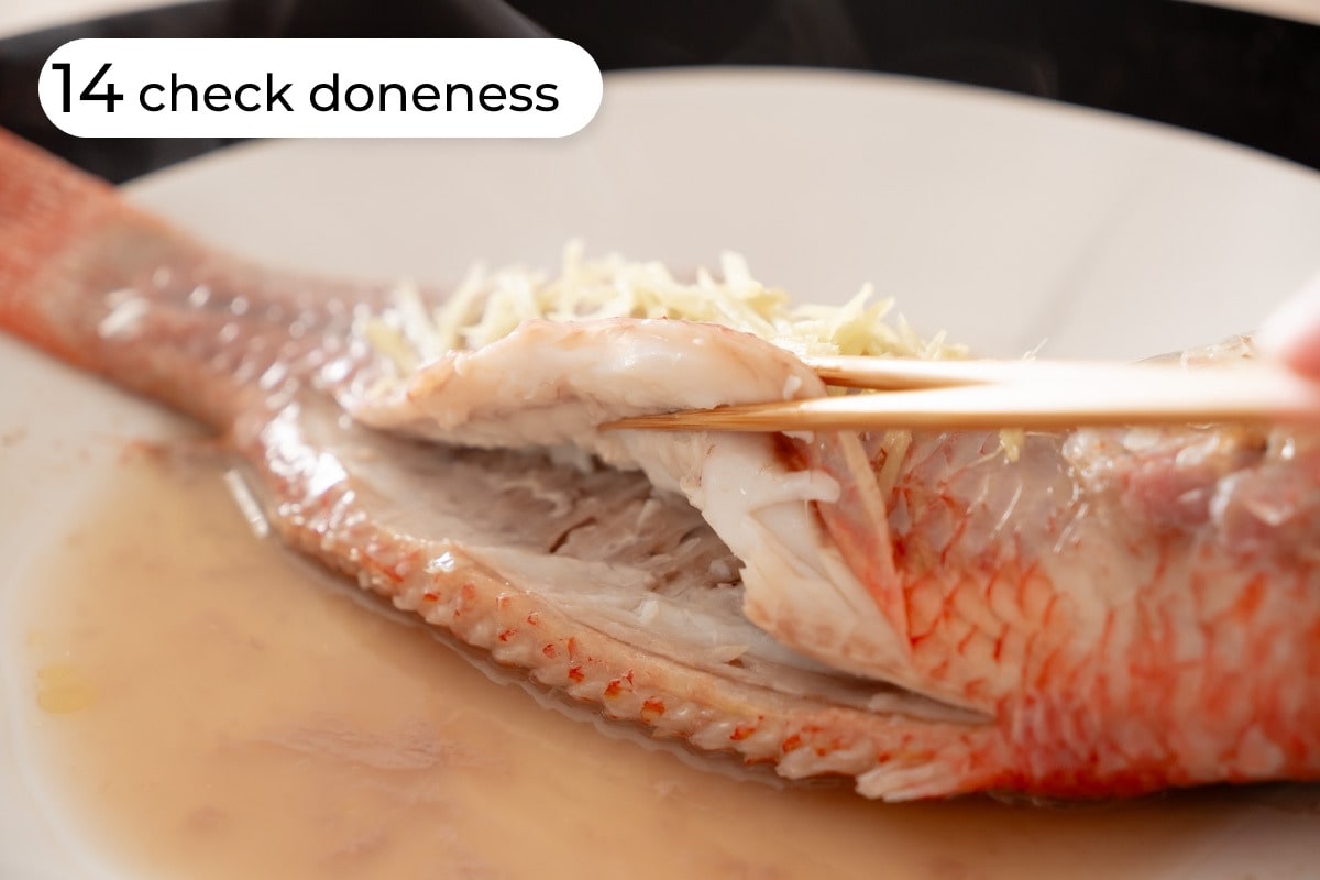 Recipe step 14 - check doneness: Freshly steamed whole fish topped with ginger slices on a plate and chopsticks pulling the meat away from the backbone to show that the meat is white and opaque while the bones are still slightly translucent, indicating the perfect doneness.