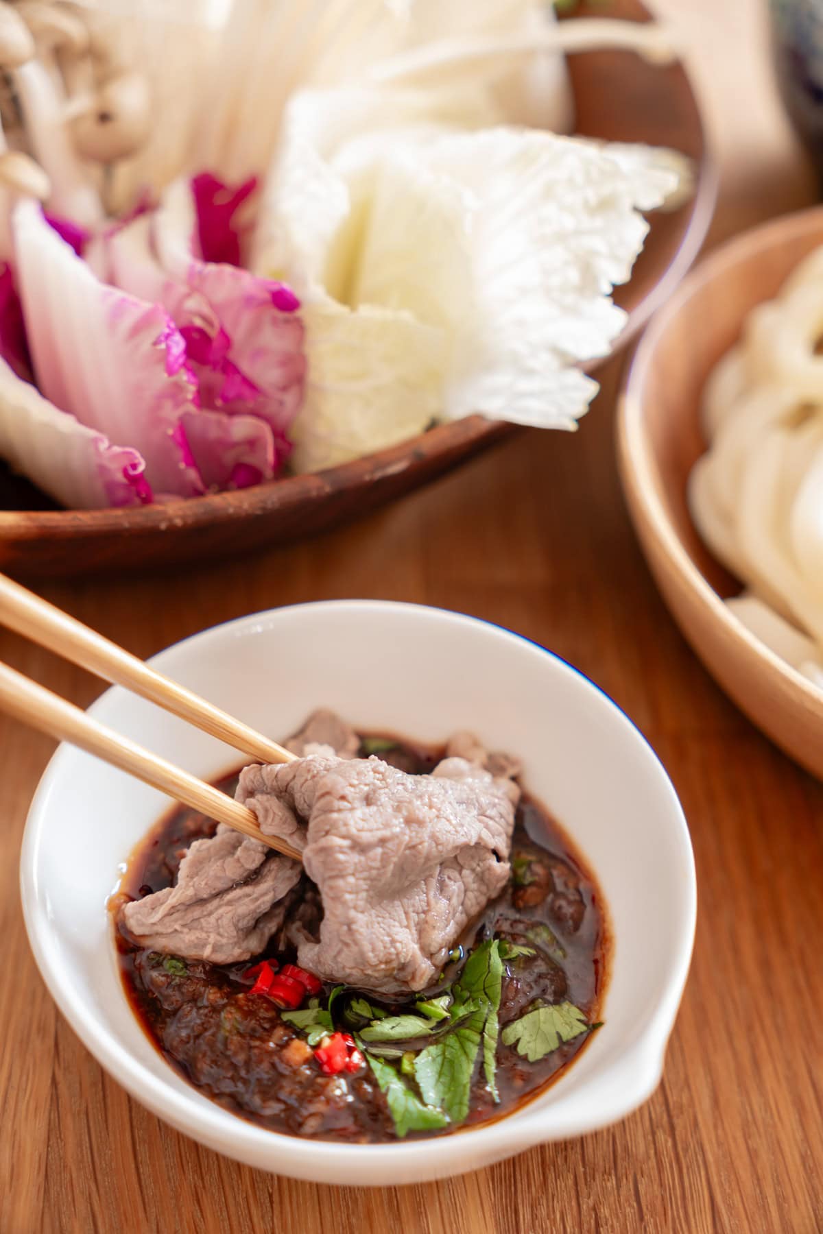 Close up view of a chopsticks holding a slice of cooked beef and dipping it in a sacha based hotpot sauce.