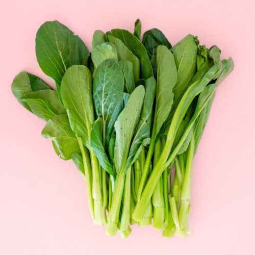 A bunch of fresh choy sum (yu choy) with broad leaves and long stems, set against a pink background.