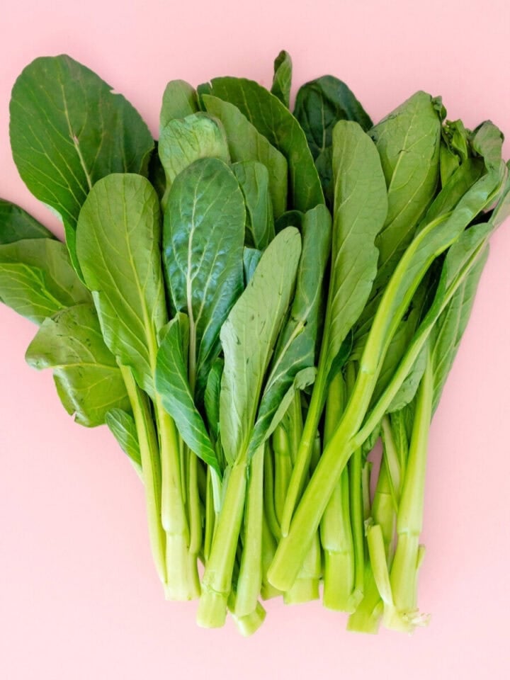 A bunch of fresh choy sum (yu choy) with broad leaves and long stems, set against a pink background.