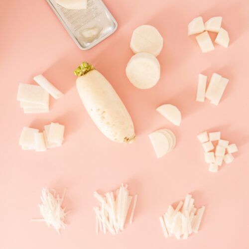 An assortment of white daikon radish pieces arranged on a pink surface. Various cuts include slices, cubes, and matchsticks, surrounding a whole radish with green leaves on top.
