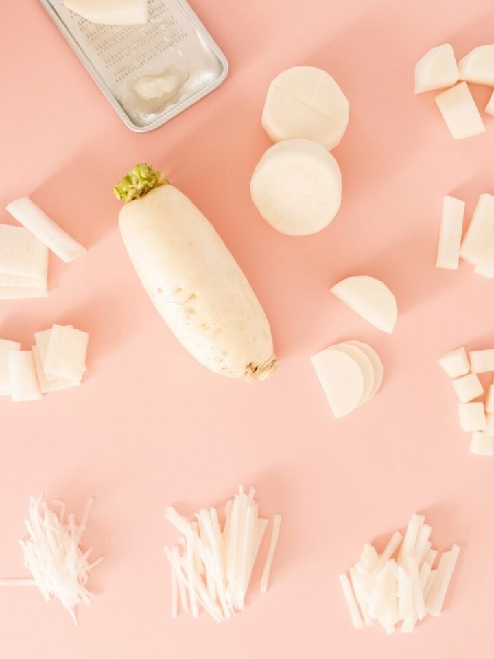 An assortment of white daikon radish pieces arranged on a pink surface. Various cuts include slices, cubes, and matchsticks, surrounding a whole radish with green leaves on top.