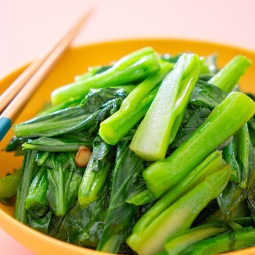 A shallow yellow bowl filled with stir-fried yu choy with garlic, accompanied by chopsticks on the side.