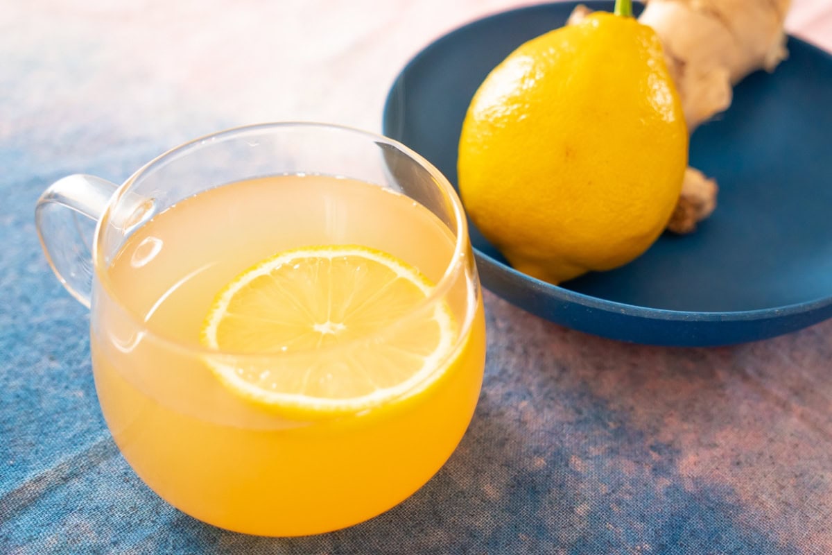 A clear glass mug filled with ginger tea with honey and a thin lemon slice on a blue and pink surface. A whole lemon and fresh ginger is in a shallow blue plate next to it.