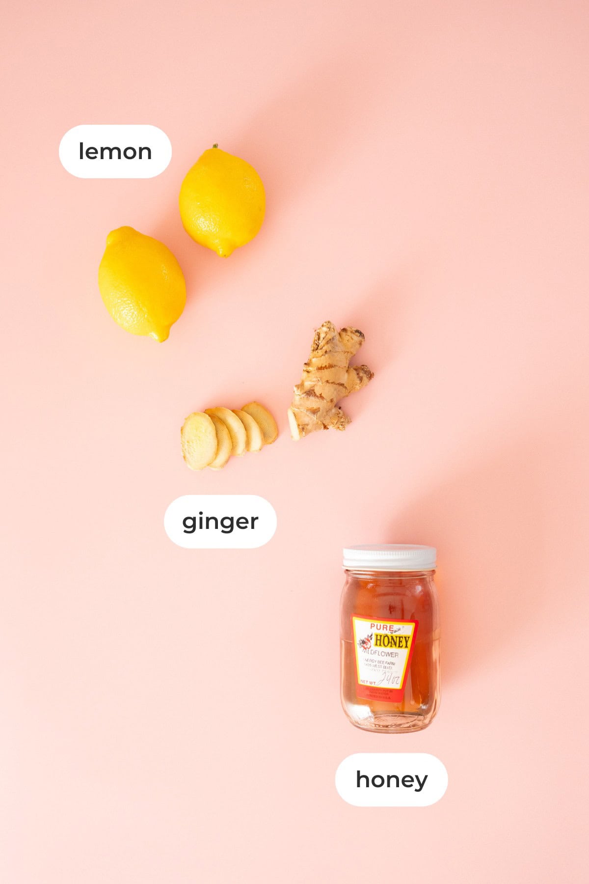 Two lemons, a piece of fresh ginger and slices, and a jar of honey arranged on a peach-colored background, each labeled accordingly.