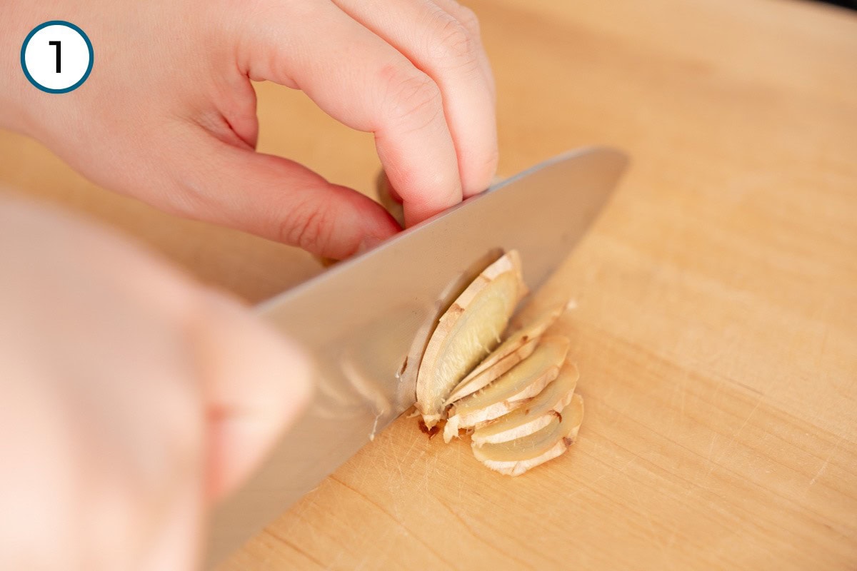 Recipe Step 1 – Close-up of Cindy slicing ginger on a wooden cutting board with a chef's knife, marked with a number 1 in the top left corner.