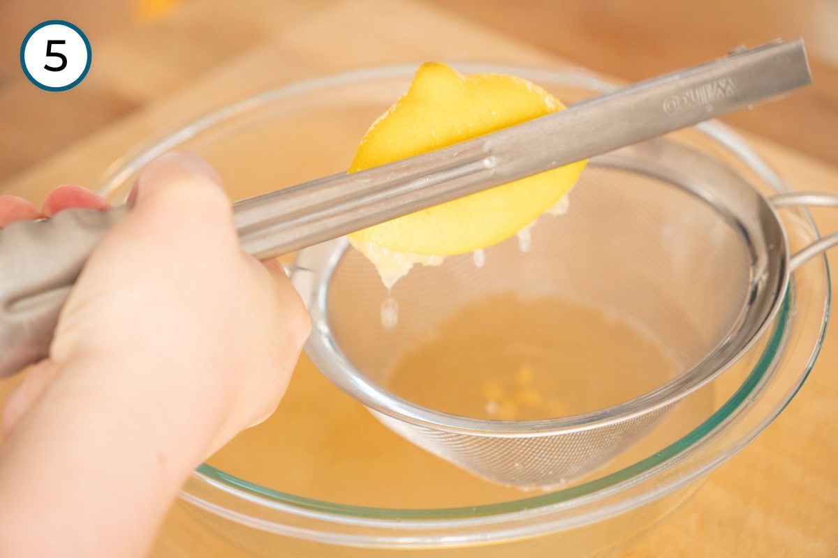 Recipe Step 5 – Cindy juicing half a lemon by squeezing it with metal tongs over a fine mesh strainer into a large glass bowl.