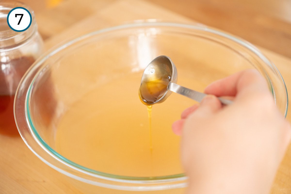 Recipe Step 7 – Cindy drizzles honey from a tablespoon measuring spoon into a large glass bowl filled with a light amber ginger tea with lemon. A honey jar is partially visible in the background.