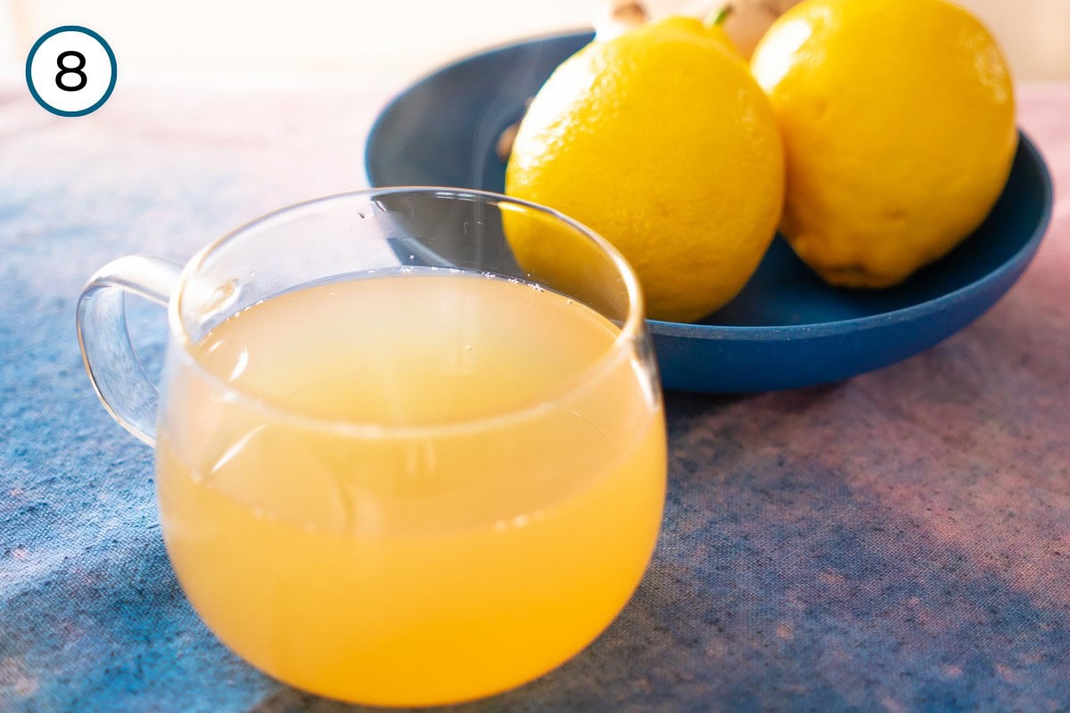 A glass mug filled with ginger lemon honey tea with steam rising, and two lemons in a dark blue shallow bowl in the background.