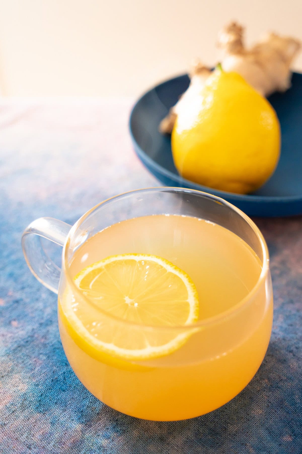 A clear glass mug filled with ginger lemon honey tea and a thin lemon slice on a blue and pink surface. In the background, a whole lemon and fresh ginger in a shallow blue plate.