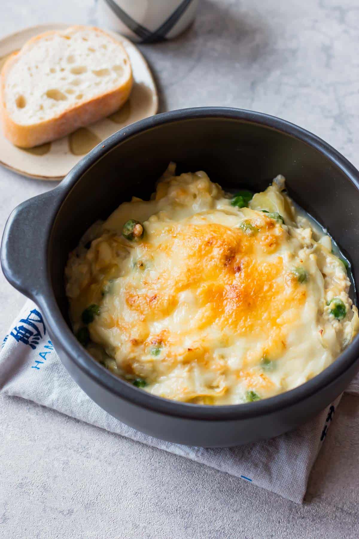 A black bowl filled with baked cheesy casserole with vegetables, sitting on a napkin next to a slice of bread on a plate.