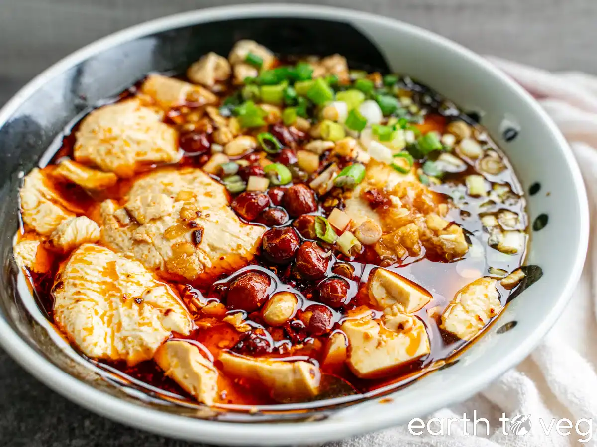 A bowl of silken tofu in Sichuan doufunao (savory tofu pudding) with chili oil, chopped green onions, and sauce, garnished with peanuts.