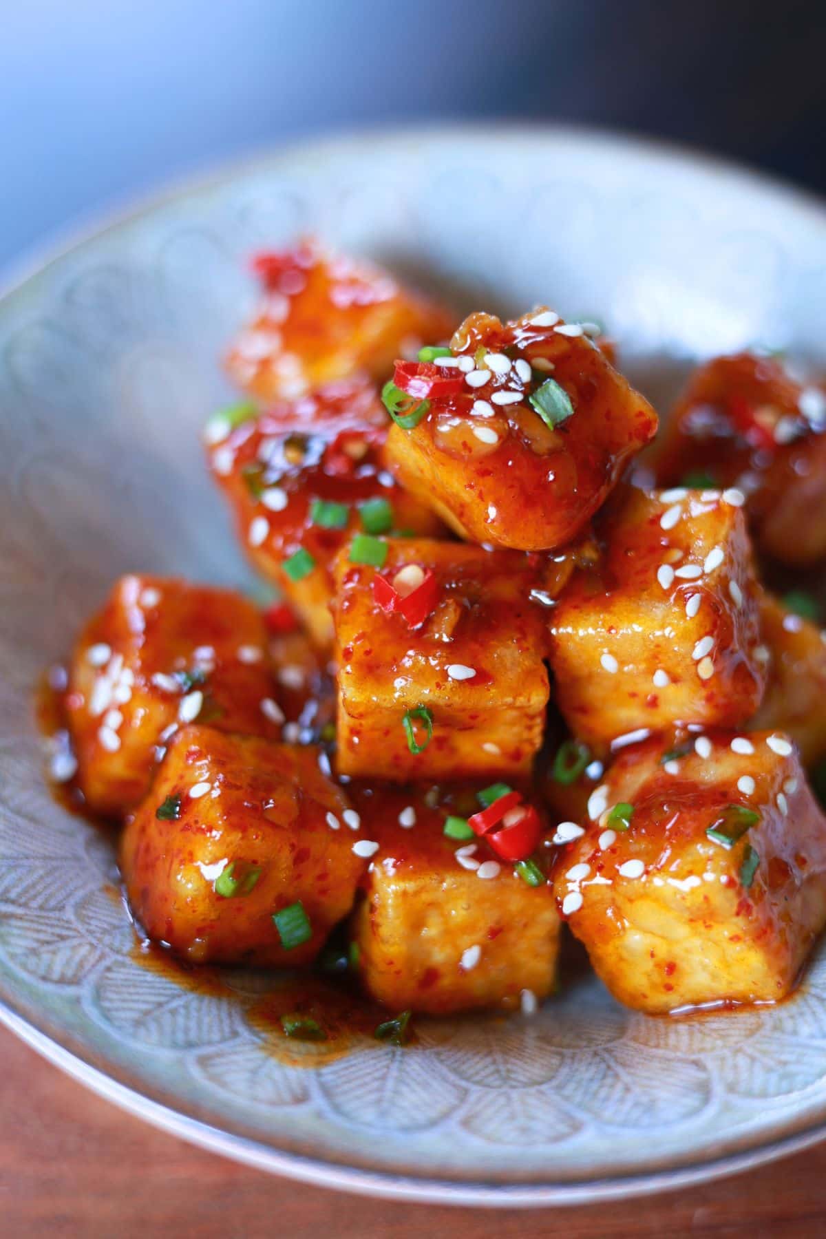 A plate of cubed tofu coated in a red, sweet and spicy sauce, garnished with chopped green onions, red chili peppers, and white sesame seeds.