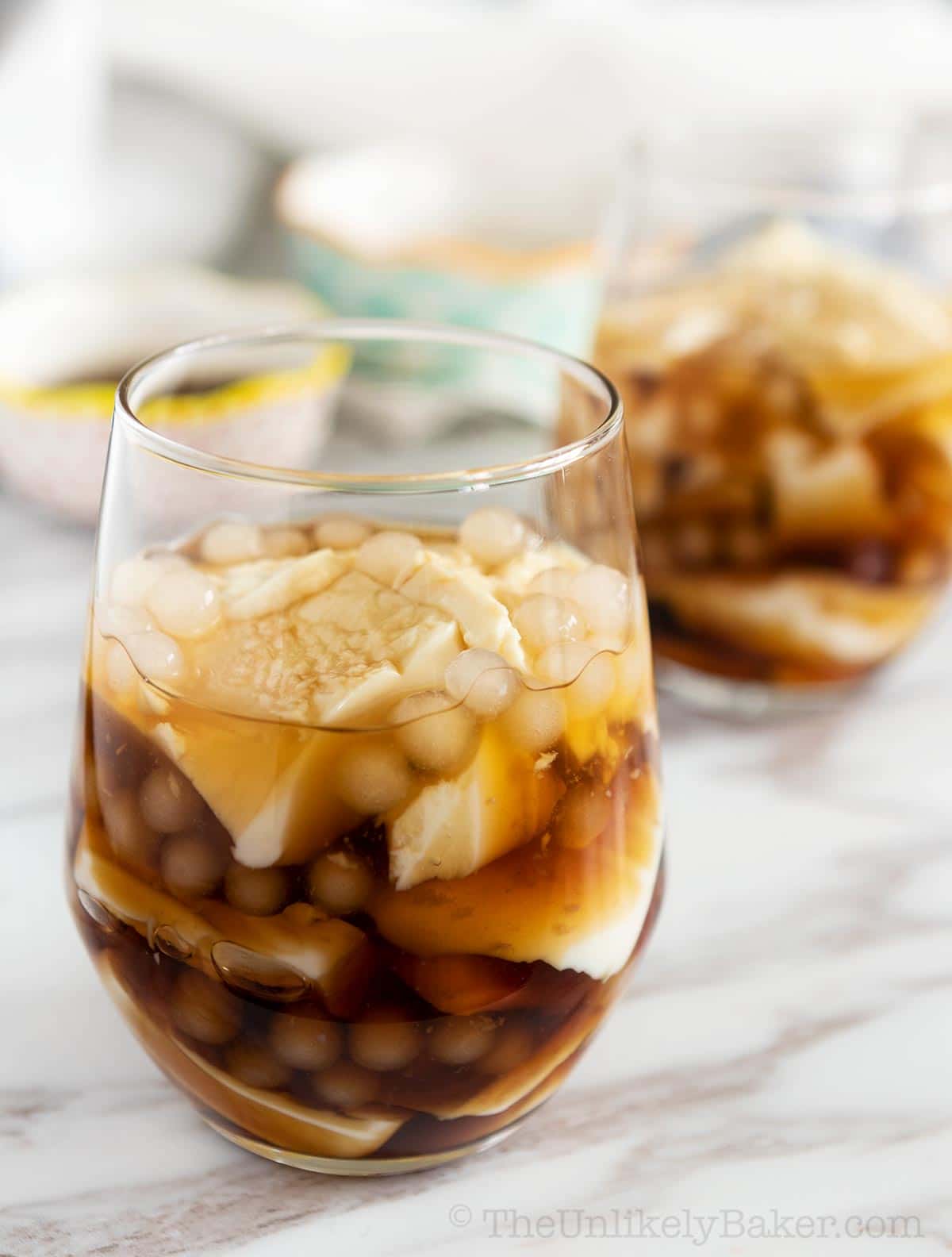 Two glasses filled with taho (Filipino sweet tofu pudding), featuring layers of silken tofu, sago pearls, and brown sugar syrup on a marble surface.