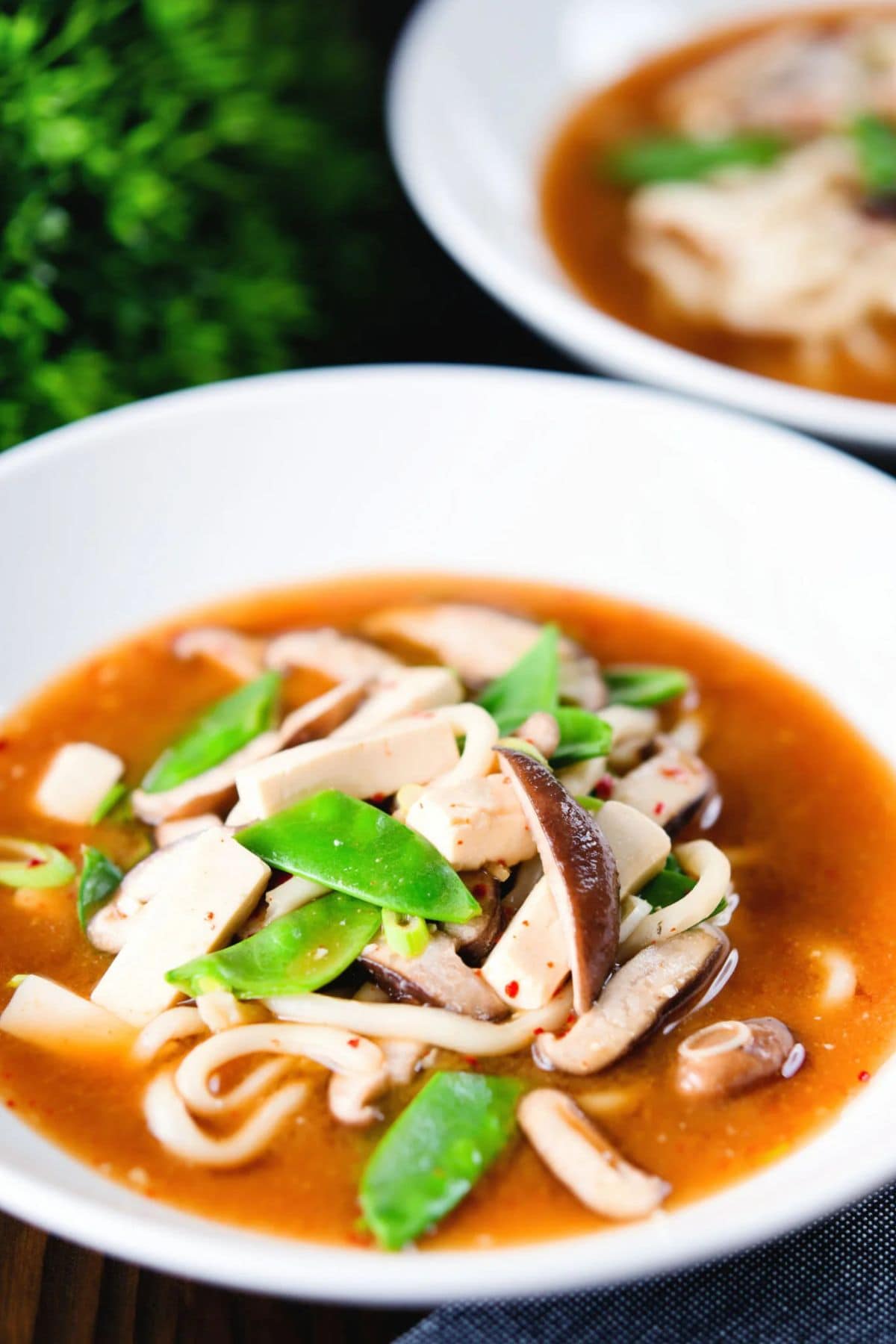 A bowl of udon noodle soup with sliced mushrooms, tofu, and snow peas in a reddish broth, with another similar bowl in the background.