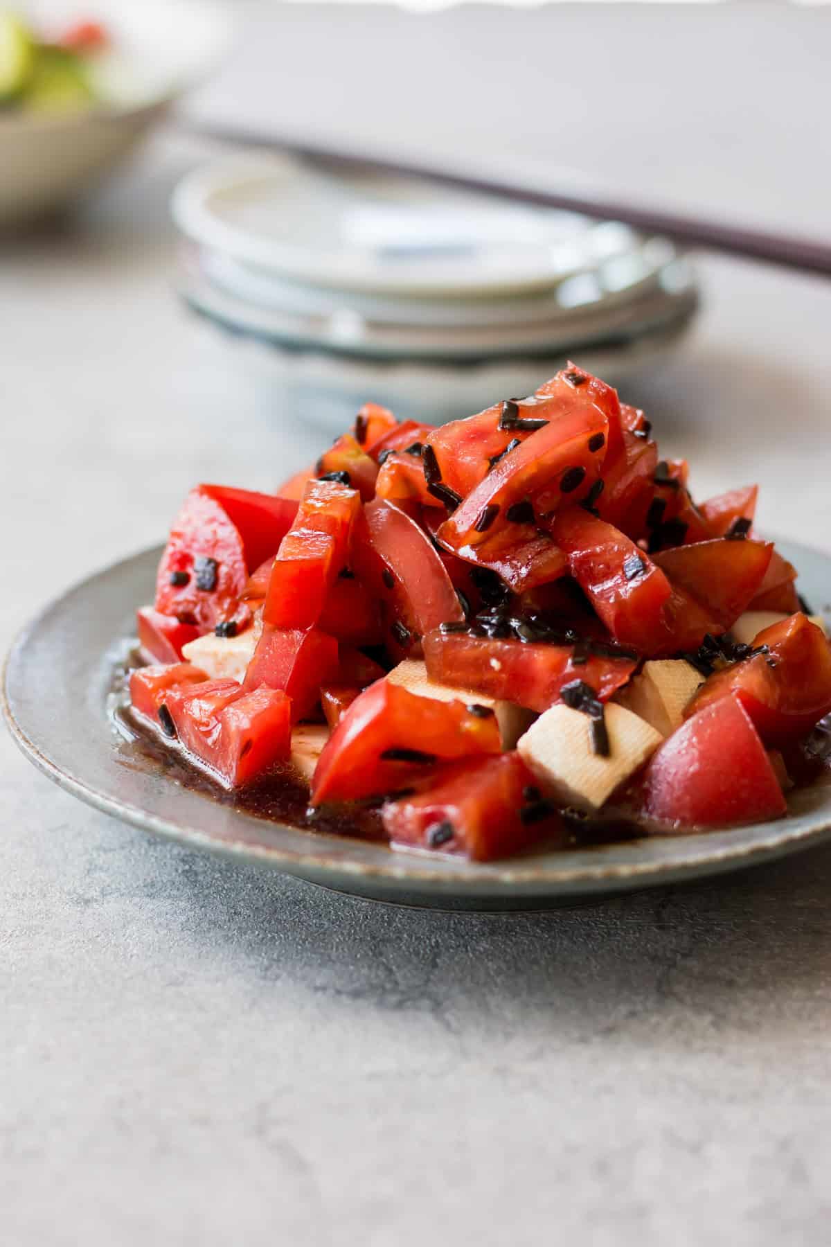 A plate of chopped tomatoes and tofu topped with seaweed and sauce, set on a gray surface with stacked plates in the background.