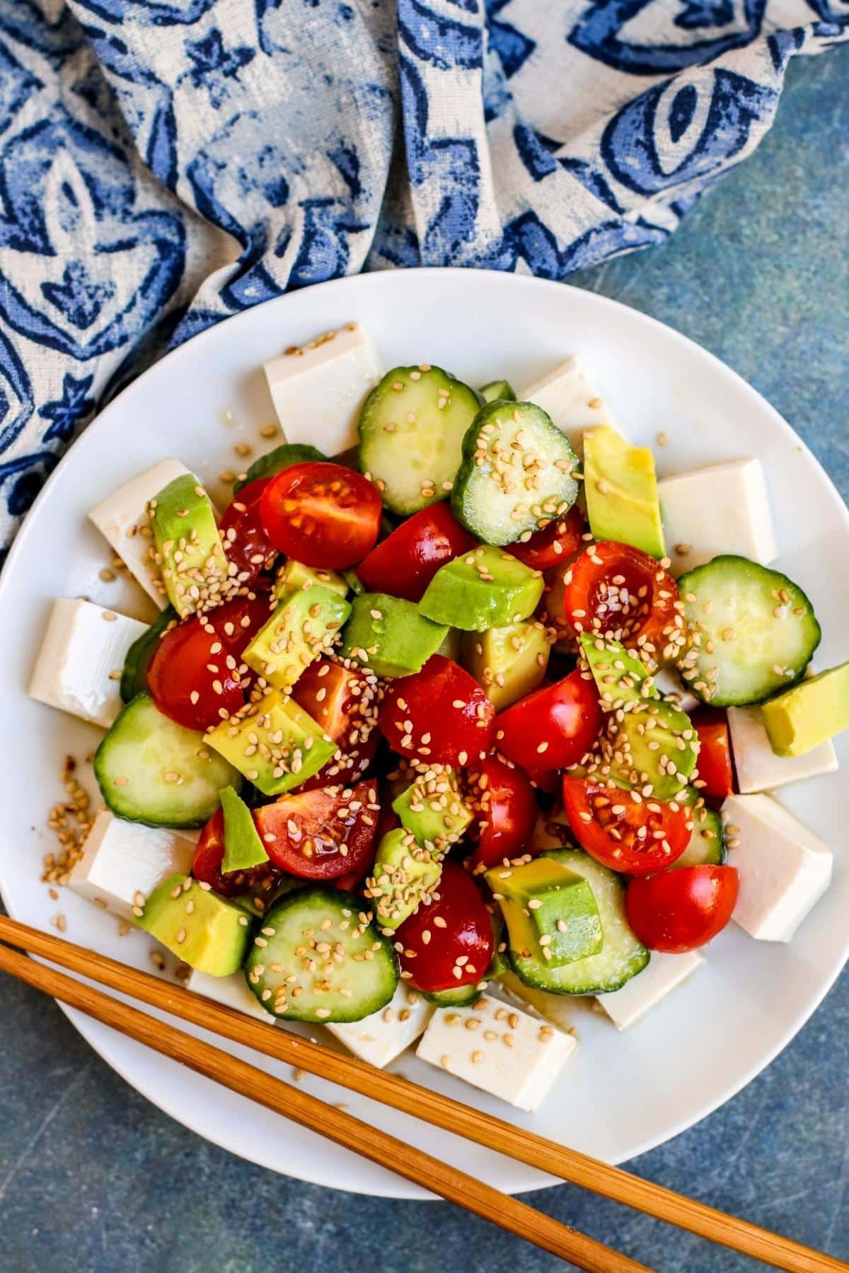 A plate of silken tofu cubes topped with sliced cucumbers, cherry tomatoes, avocado, and sprinkled with sesame seeds with a dressing; chopsticks rest on the plate.