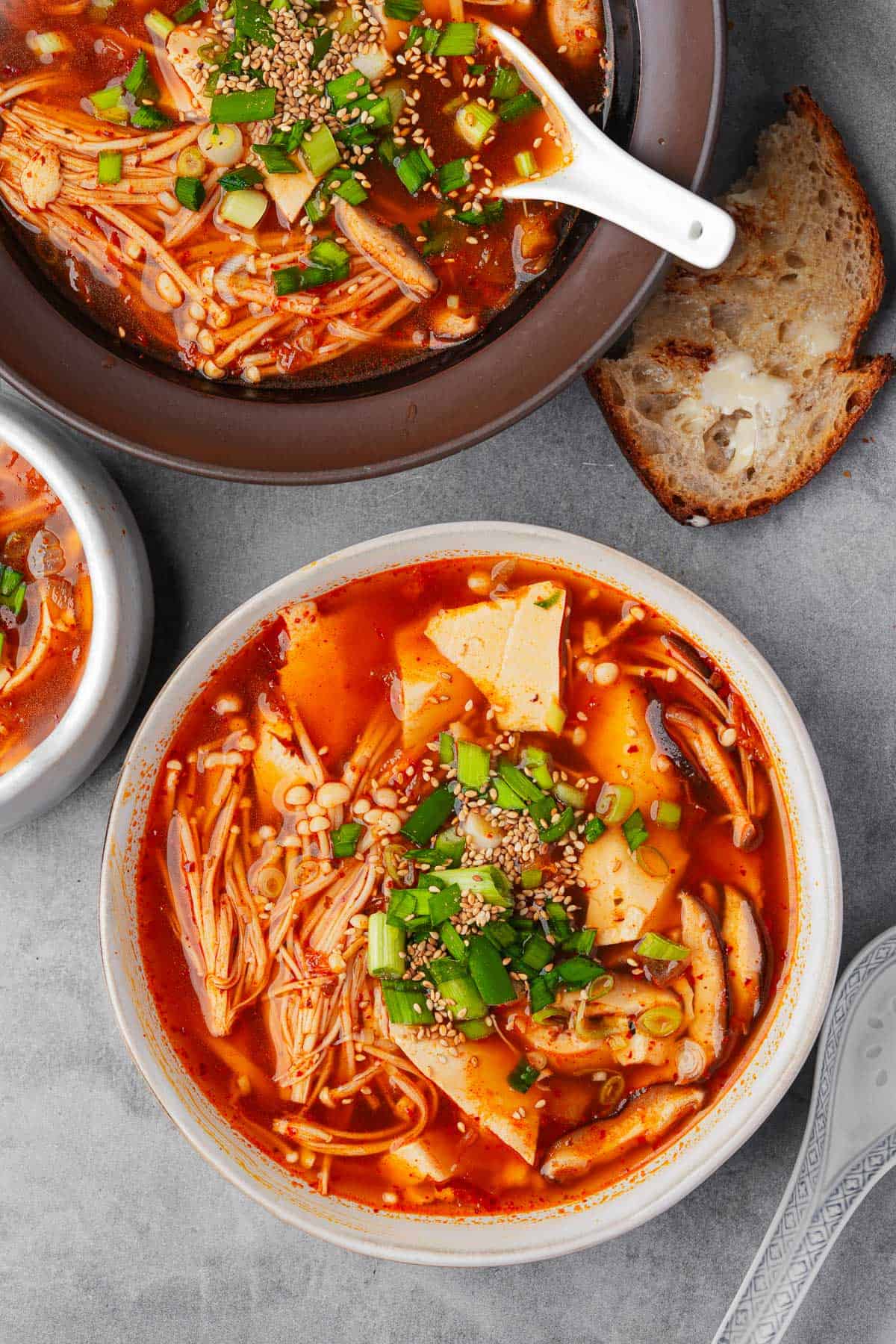 Top-down view of two bowls of spicy sundubu jjigae (Korean tofu soup), mushrooms, green onions, and sesame seeds on a gray surface.