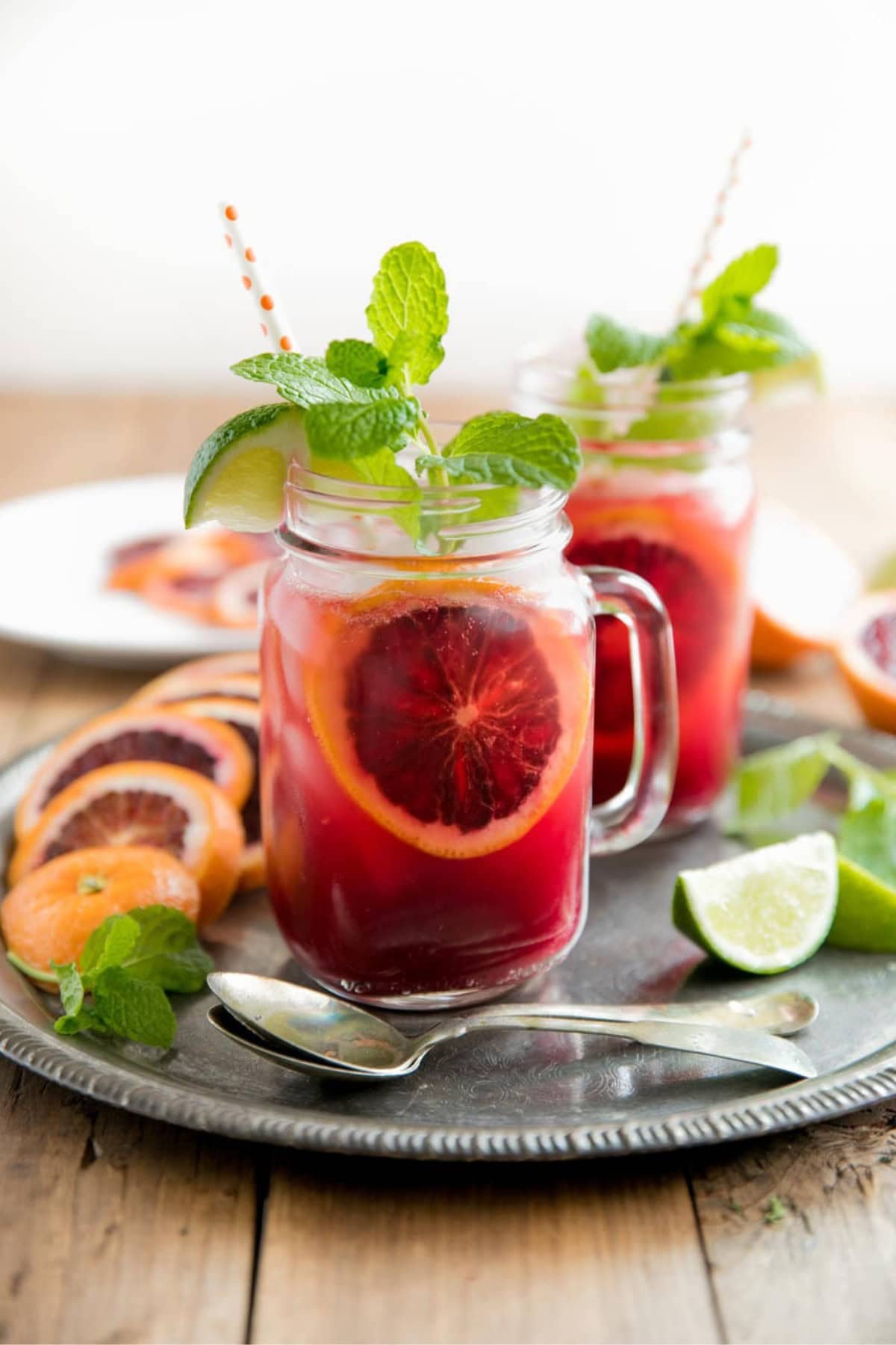 Two mason jars filled with a red citrus drink, garnished with blood orange slices, mint sprigs, and lime wedges, placed on a metal tray with fruit and a spoon.