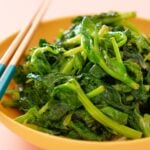 Stir-fried pea shoots in a shallow yellow serving bowl with a pair of chopsticks resting on the edge of the bowl.