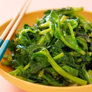 Stir-fried pea shoots in a shallow yellow serving bowl with a pair of chopsticks resting on the edge of the bowl.