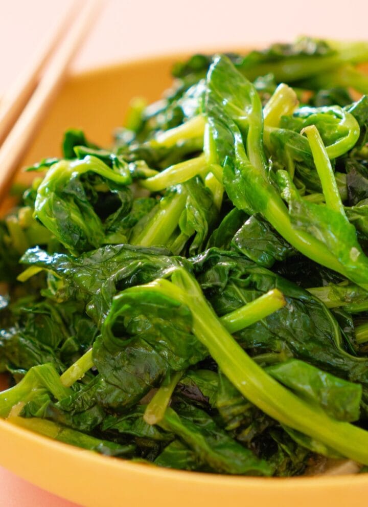 Stir-fried pea shoots in a shallow yellow serving bowl with a pair of chopsticks resting on the edge of the bowl.