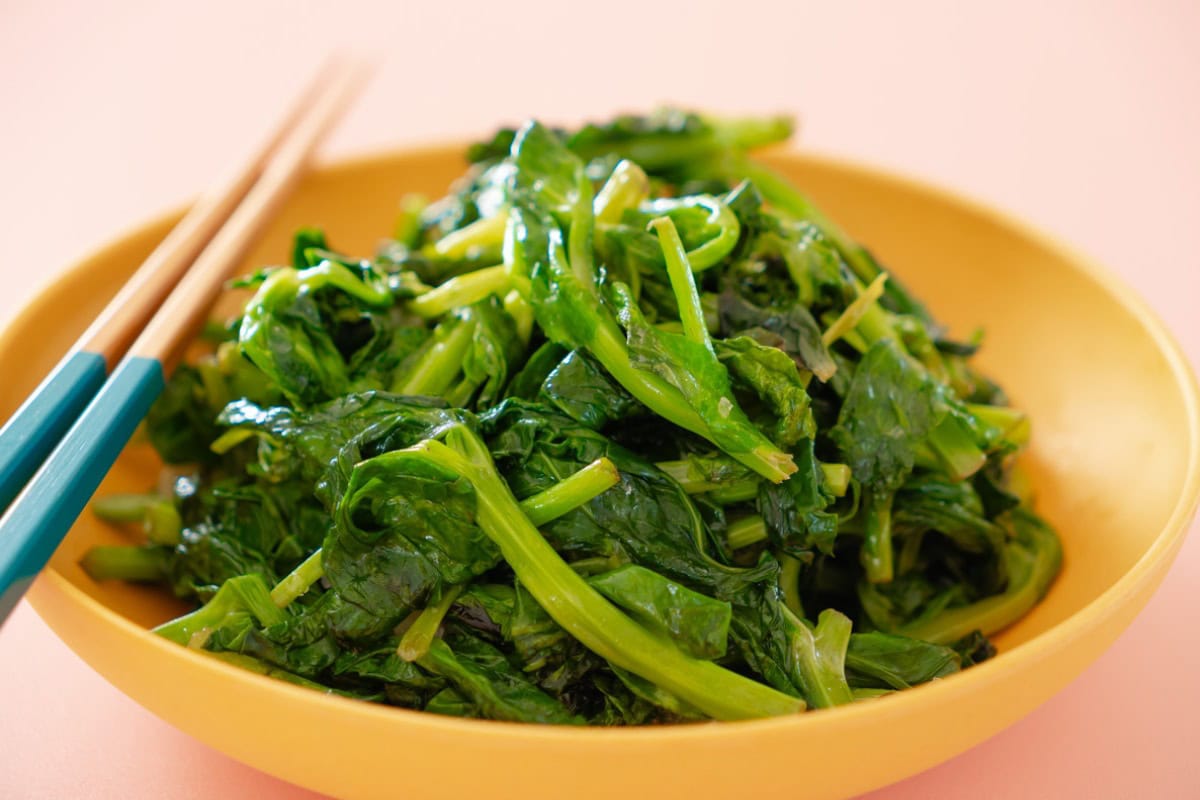 Stir-fried pea shoots in a shallow yellow serving bowl with a pair of chopsticks resting on the edge of the bowl, set against a light pink background.